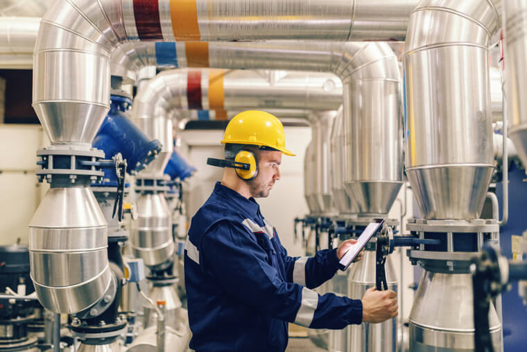 Profile of young Caucasian worker tightening the valve and using tablet while standing in heating plant